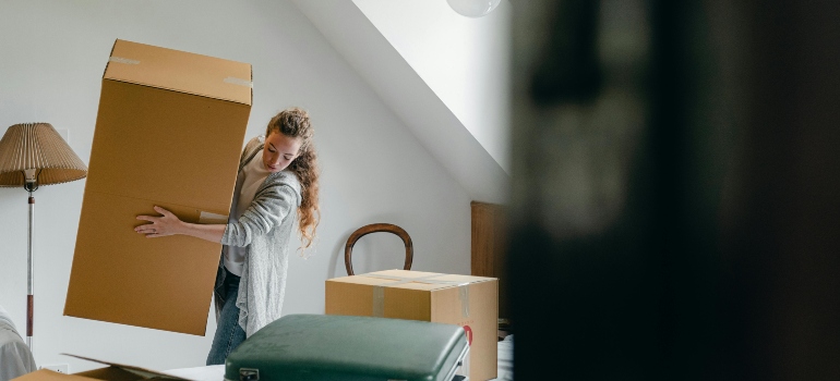 Woman lifting a cardboard box in a bright room with a lamp, chair, and suitcase.