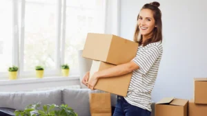 A woman stands in the living room of her new home holding a brown cardboard box full of her belongings.