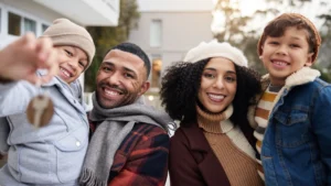 Young family wearing hats and scarves smiles and holds out keys to new home.
